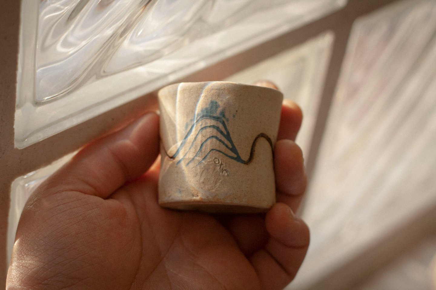 Mano sosteniendo una pequeña taza artesanal de cerámica con líneas onduladas azules y marrones.